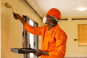 professional painter wearing an orange jumpsuit, hardhat and mask painting a home tan